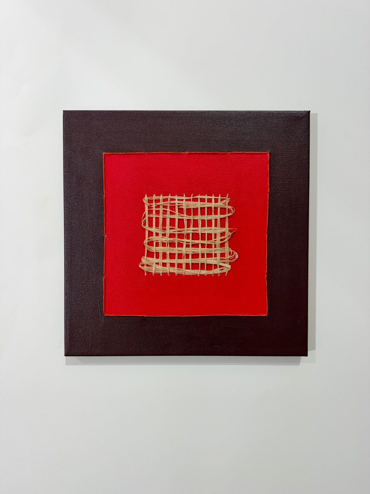A square abstract wall art piece featuring a dark brown textured frame around a bright red center panel with a beige woven geometric pattern, mounted on a light gray wall.